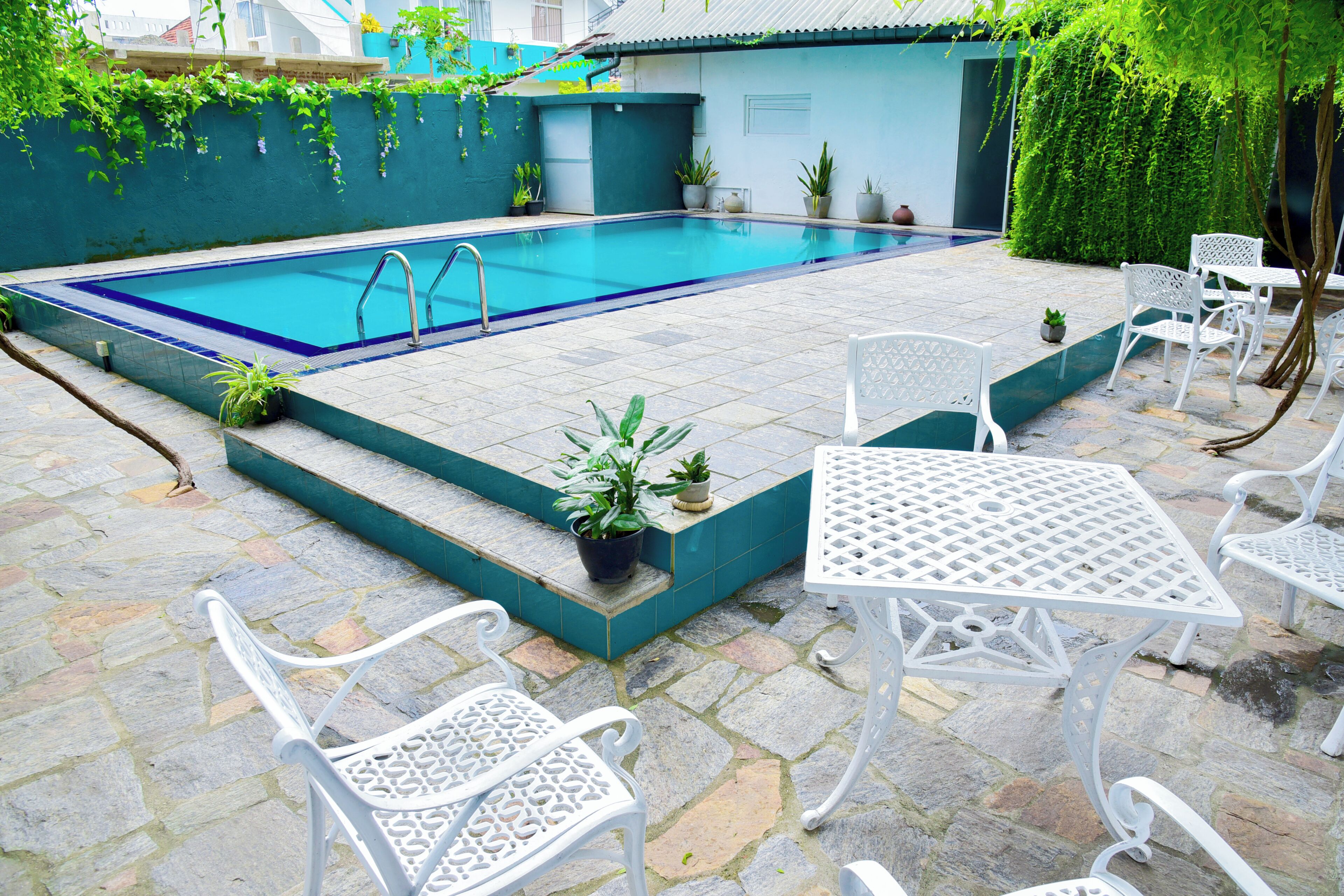 Une piscine entourée d’une terrasse carrelée, de chaises et de tables en métal blanc, et de plantes en pot.