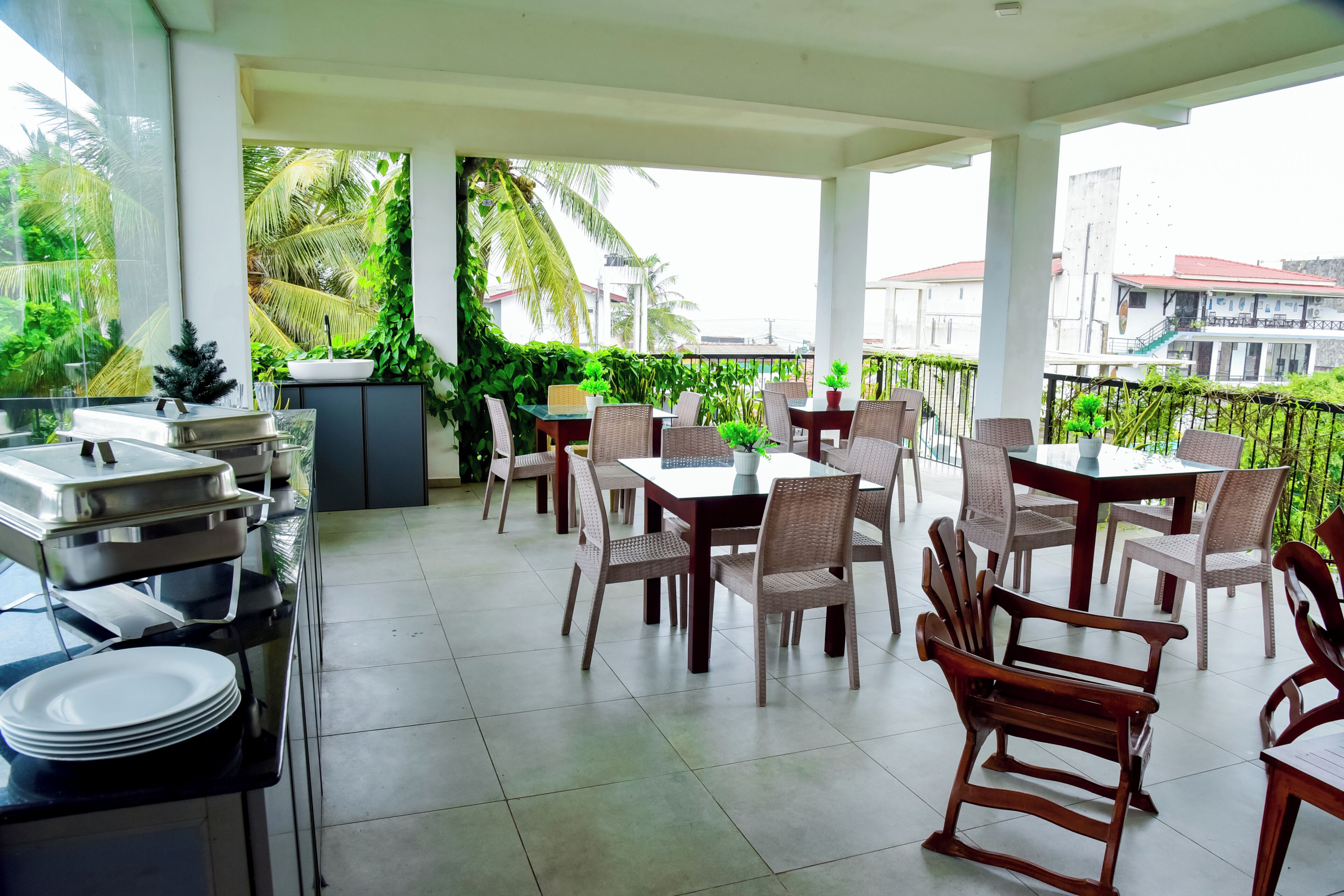 A covered outdoor dining area with tables and chairs, a buffet setup, and lush greenery visible through the glass.