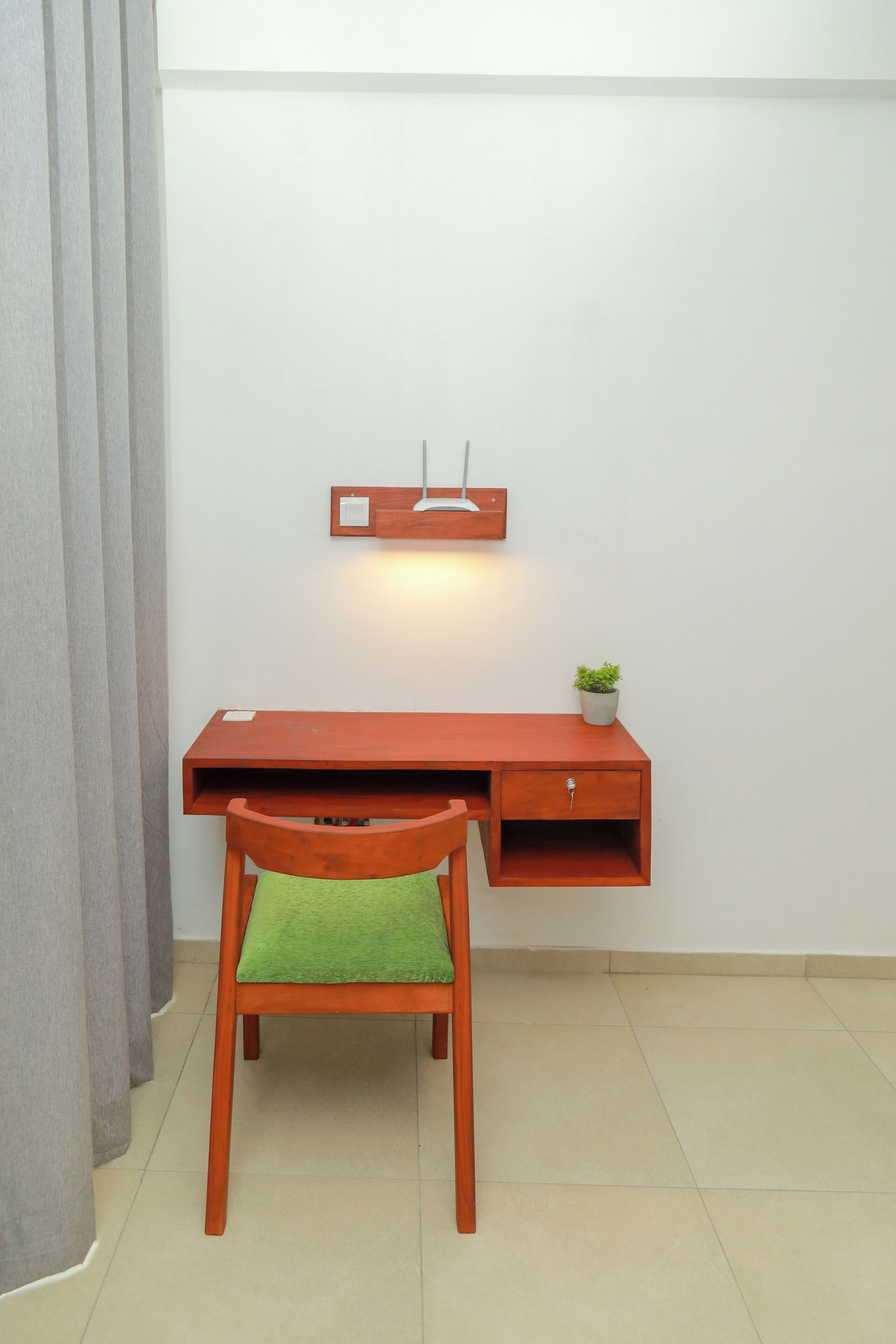 Un espacio de trabajo compacto con un escritorio de madera, una silla con cojín verde, una planta pequeña en maceta y una lámpara fijada a la pared.