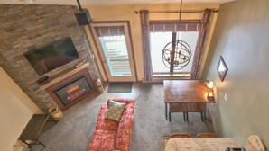 A cozy living room with a fireplace, a sofa, a dining table, and a large window with curtains. - Bayfield Condo 1BR + Loft | Lake Superior View (Bayfield)