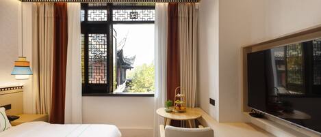 A hotel room with a bed, a TV, a small table with a plant, and a window with curtains.