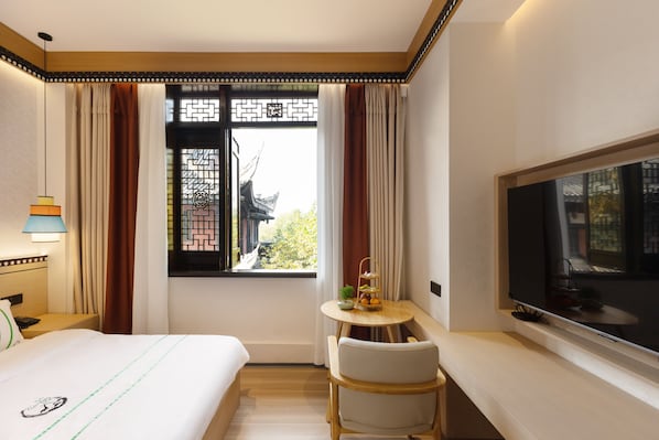 A hotel room with a bed, a TV, a small table with a plant, and a window with curtains.