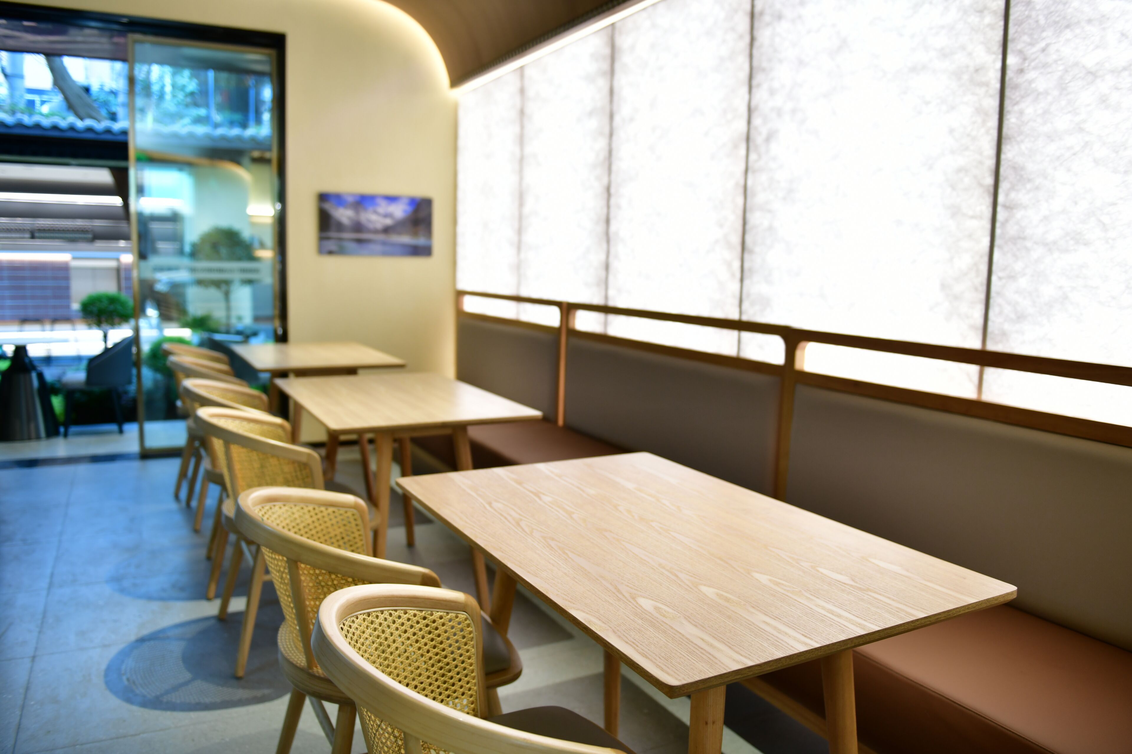 A restaurant interior with wooden tables and woven chairs, a large window with a view, and a wall-mounted picture.