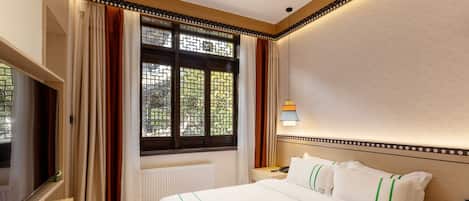 A hotel room with a large bed, a TV, a window with curtains, and a bedside table with a lamp.