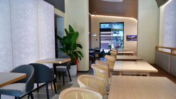 A modern restaurant interior with wooden tables and chairs, a potted plant, and a view of the kitchen through a glass wall.