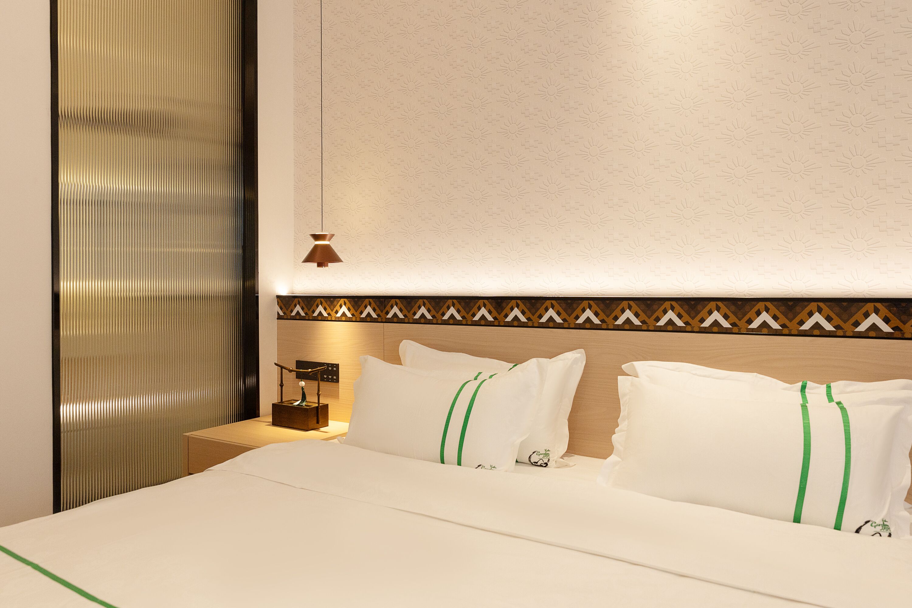 A hotel room with a neatly made bed, a bedside table with a lamp, and a decorative wall panel.