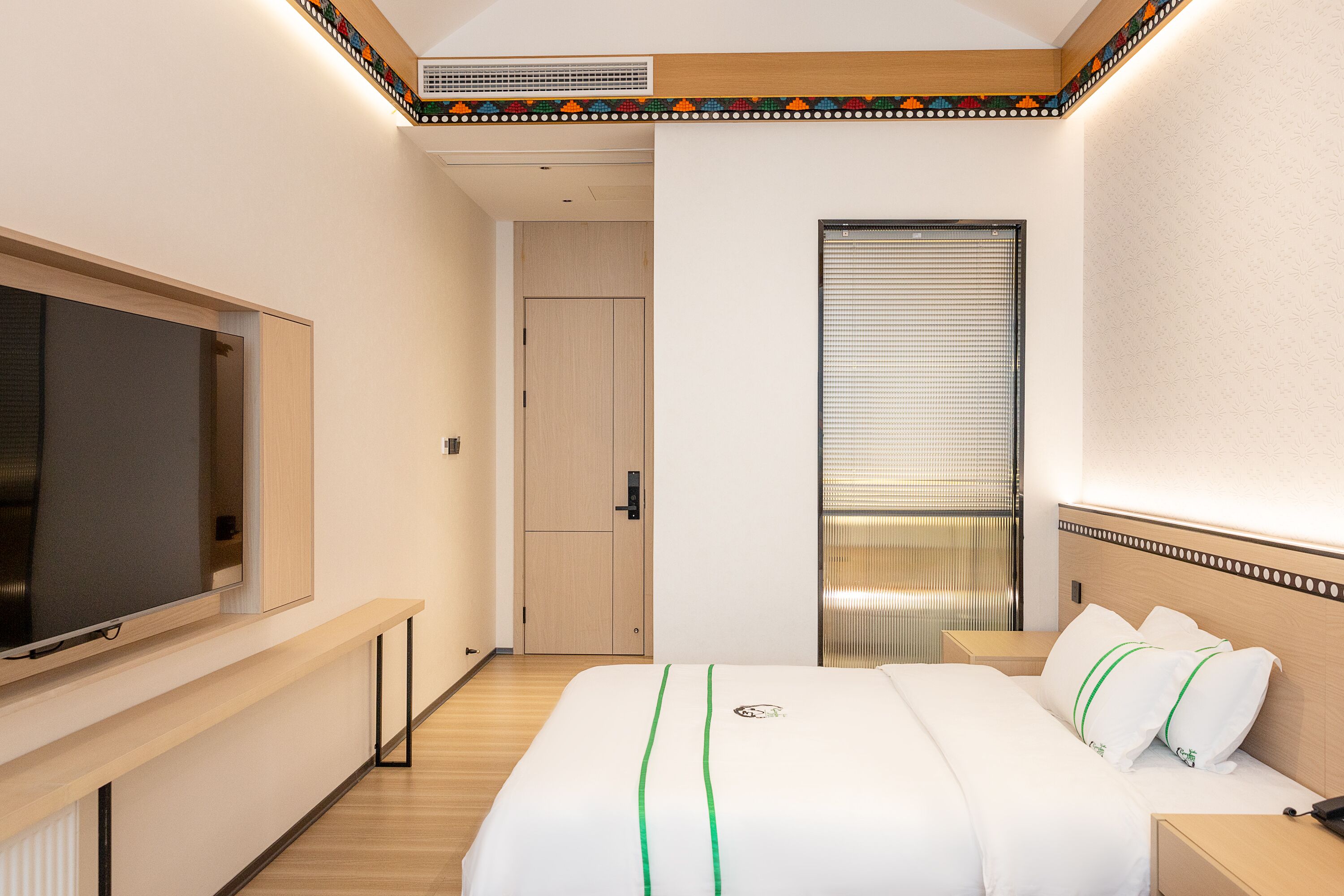 A hotel room with a bed, a TV, a window with blinds, and a door leading to another room.