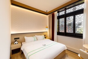 A hotel room with a neatly made bed, a bedside table with a lamp, a nightstand with a panda-shaped alarm clock, and a window with decorative glass panes.