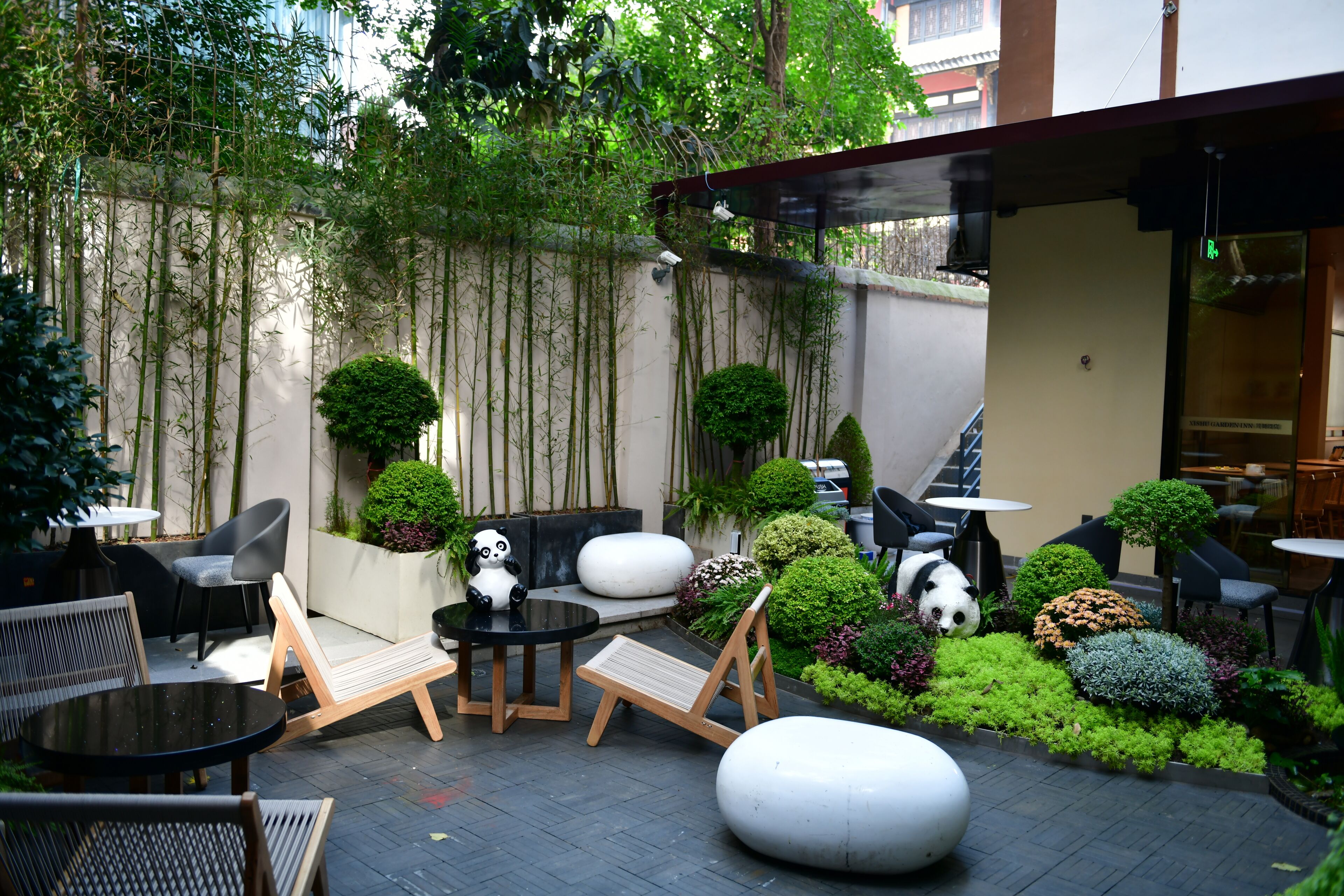 Un espacio de patio con mobiliario exterior, incluyendo sillas y mesas, rodeado de plantas variadas y una estatua de panda.