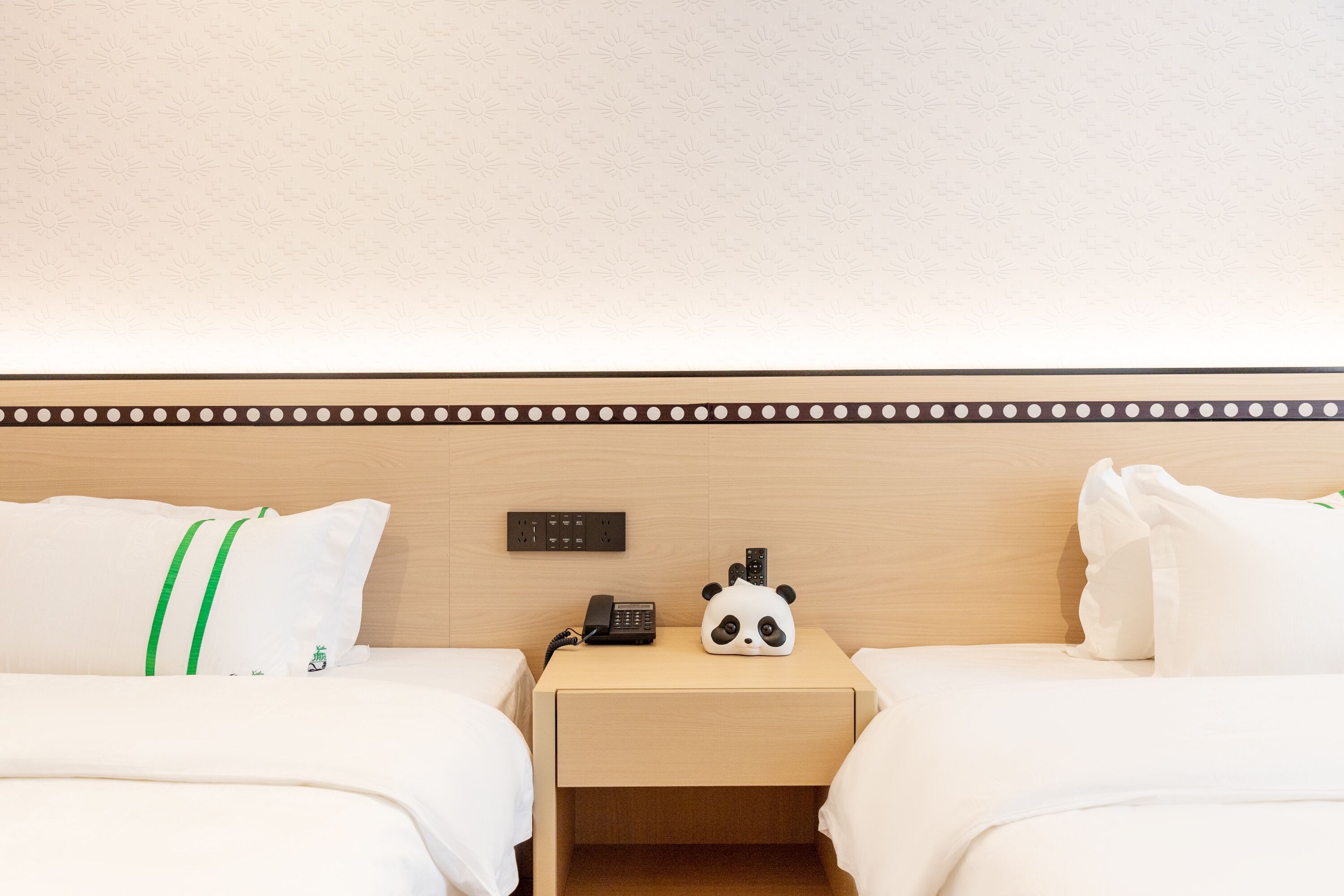 A hotel room with two beds, a nightstand with a phone and a panda-shaped lamp, and a wooden headboard with a decorative border.