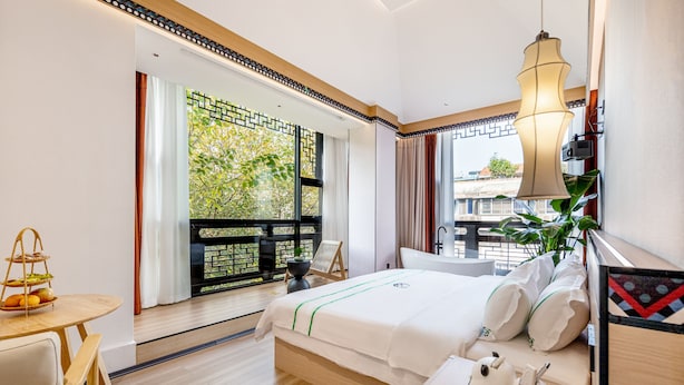 A modern hotel room with a large bed, a small table with a basket of fruit, a view of greenery outside, and a balcony with a railing. - XISHU GARDEN INN (Chengdu)