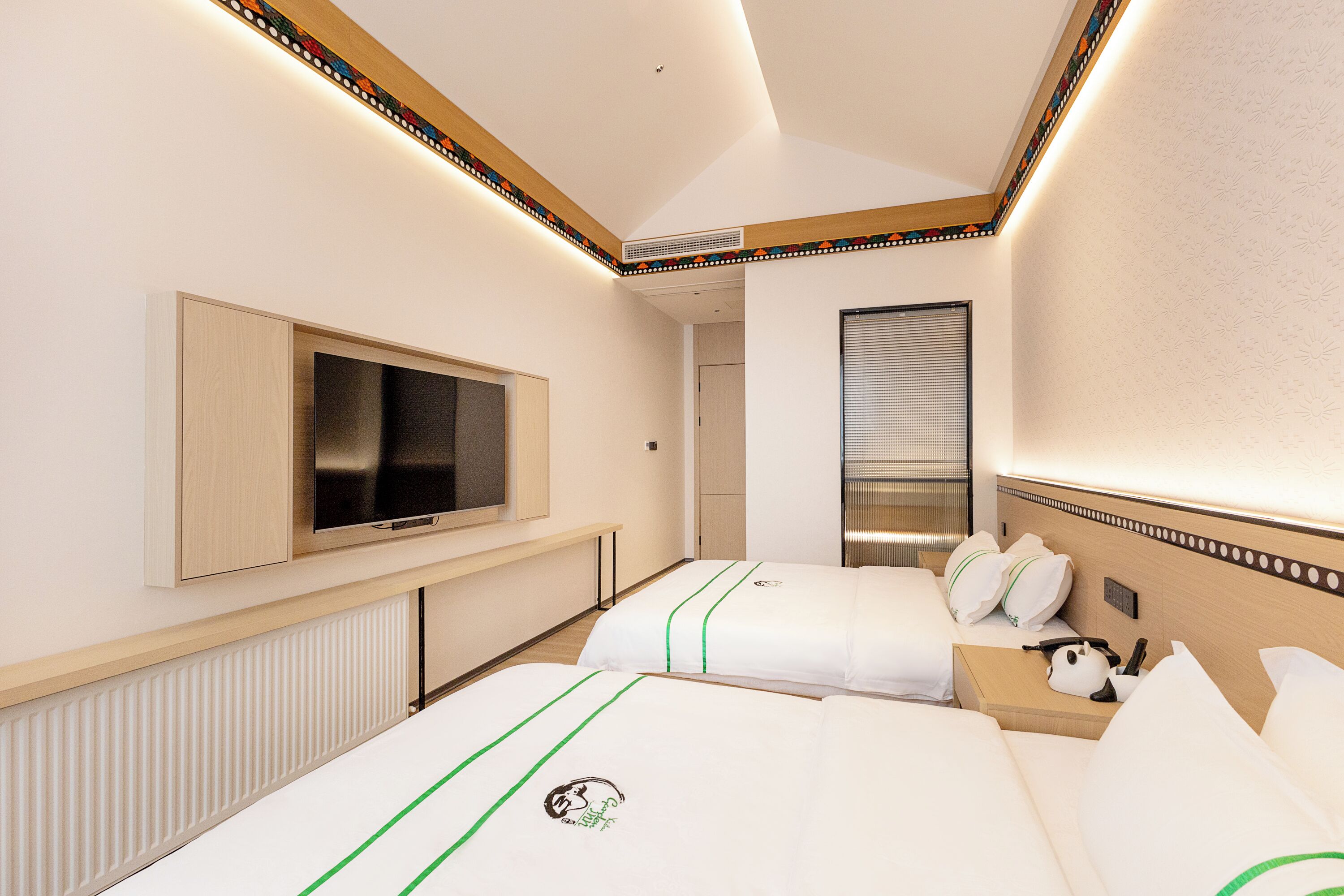 A hotel room with two beds, a TV, a window with blinds, and a bedside table with a lamp.