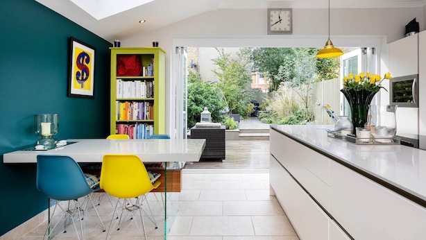Een moderne keuken met een wit aanrecht, een gele hanglamp en uitzicht op de tuin via de open deur.