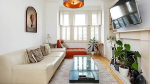 A living room with a sofa, a glass coffee table, a TV mounted on the wall, and a large window with shutters. - The Tranquil Townhouse, by Veeve (London)