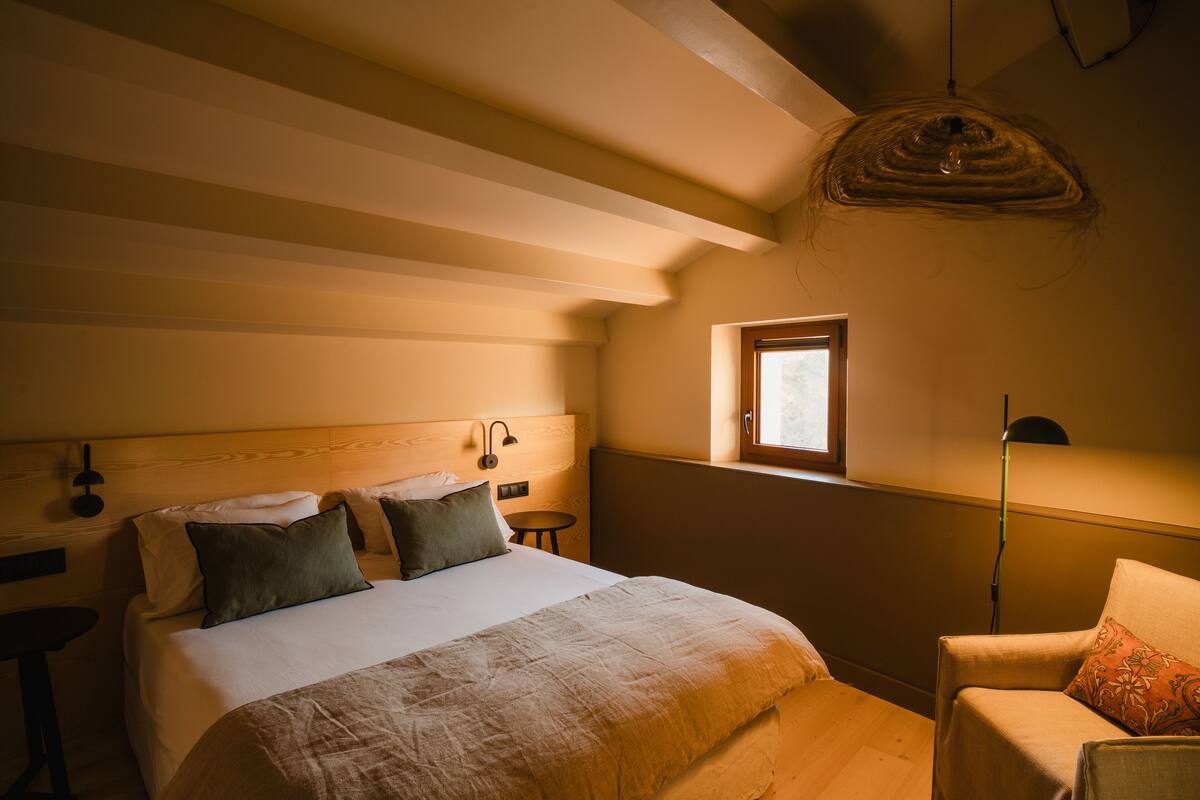 A bedroom with a bed, a chair, a small table, and a window.