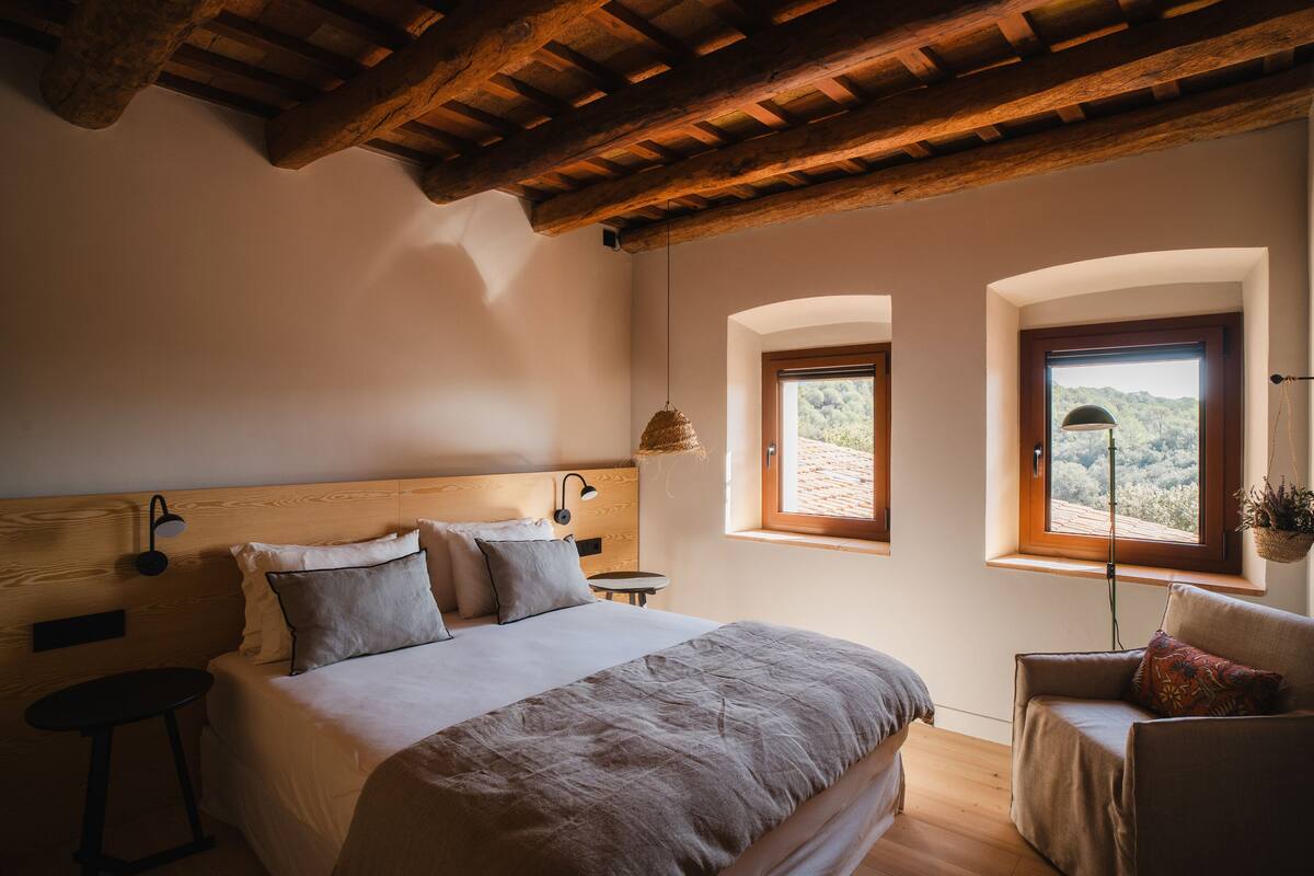 A bedroom with a wooden ceiling, a bed with a headboard, two windows, and a small table.