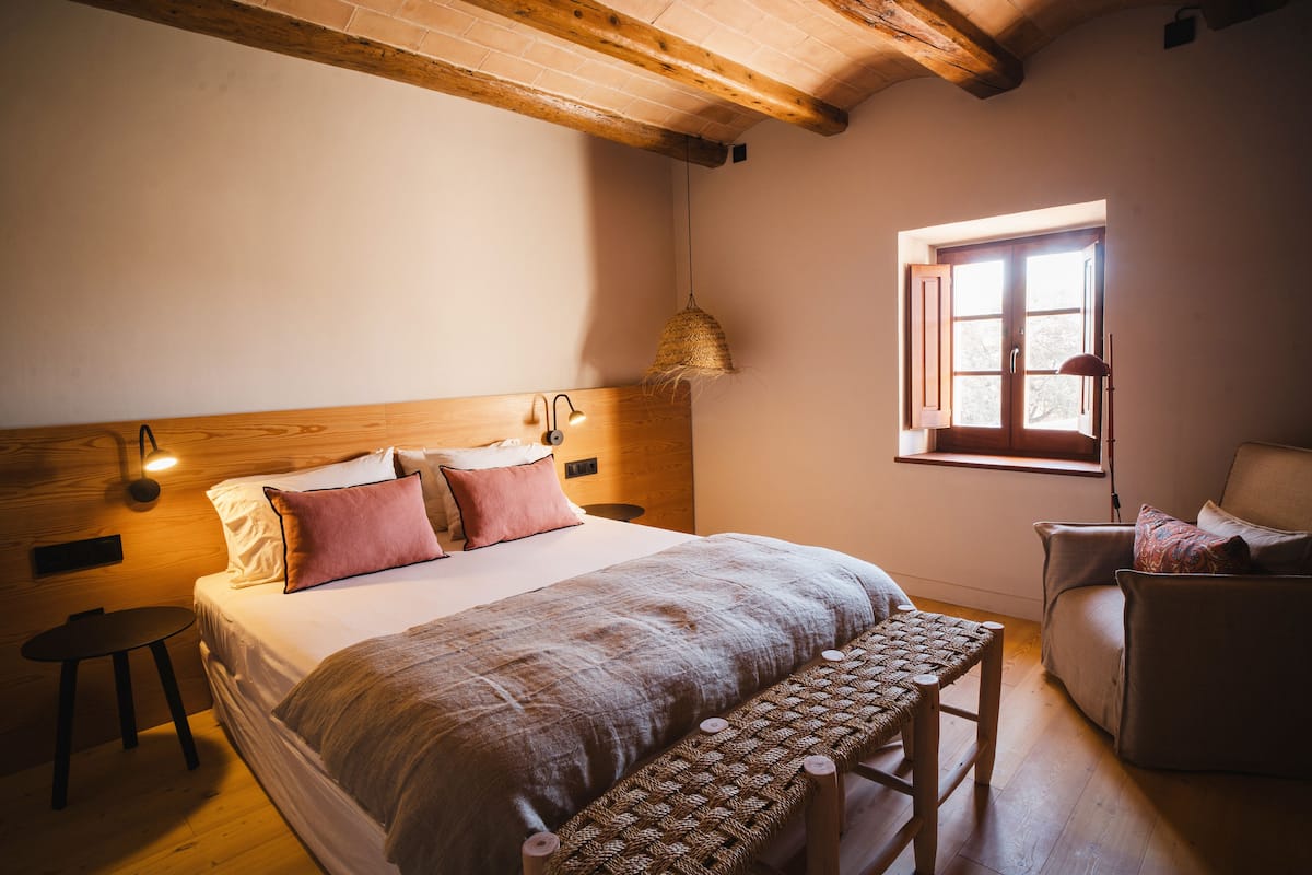 A cozy bedroom with a wooden headboard, a large bed with a patterned blanket, a small round table, and a window with a view.