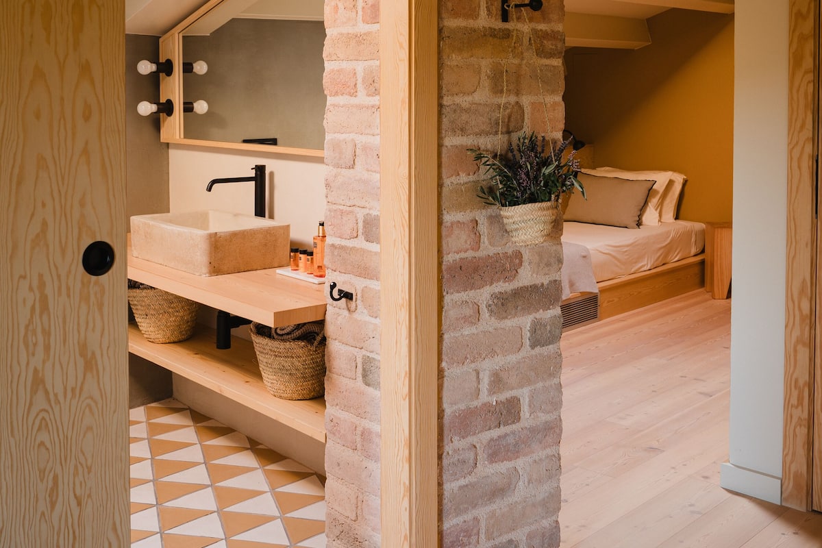 A room with a wooden floor, a brick wall, a wooden bench, a bed, and a mirror with lights.