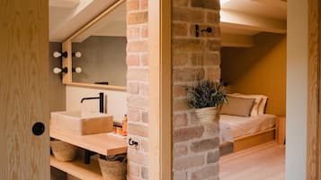 Una habitación con suelo de madera, pared de ladrillo, un banco de madera, una cama y un espejo con luces.