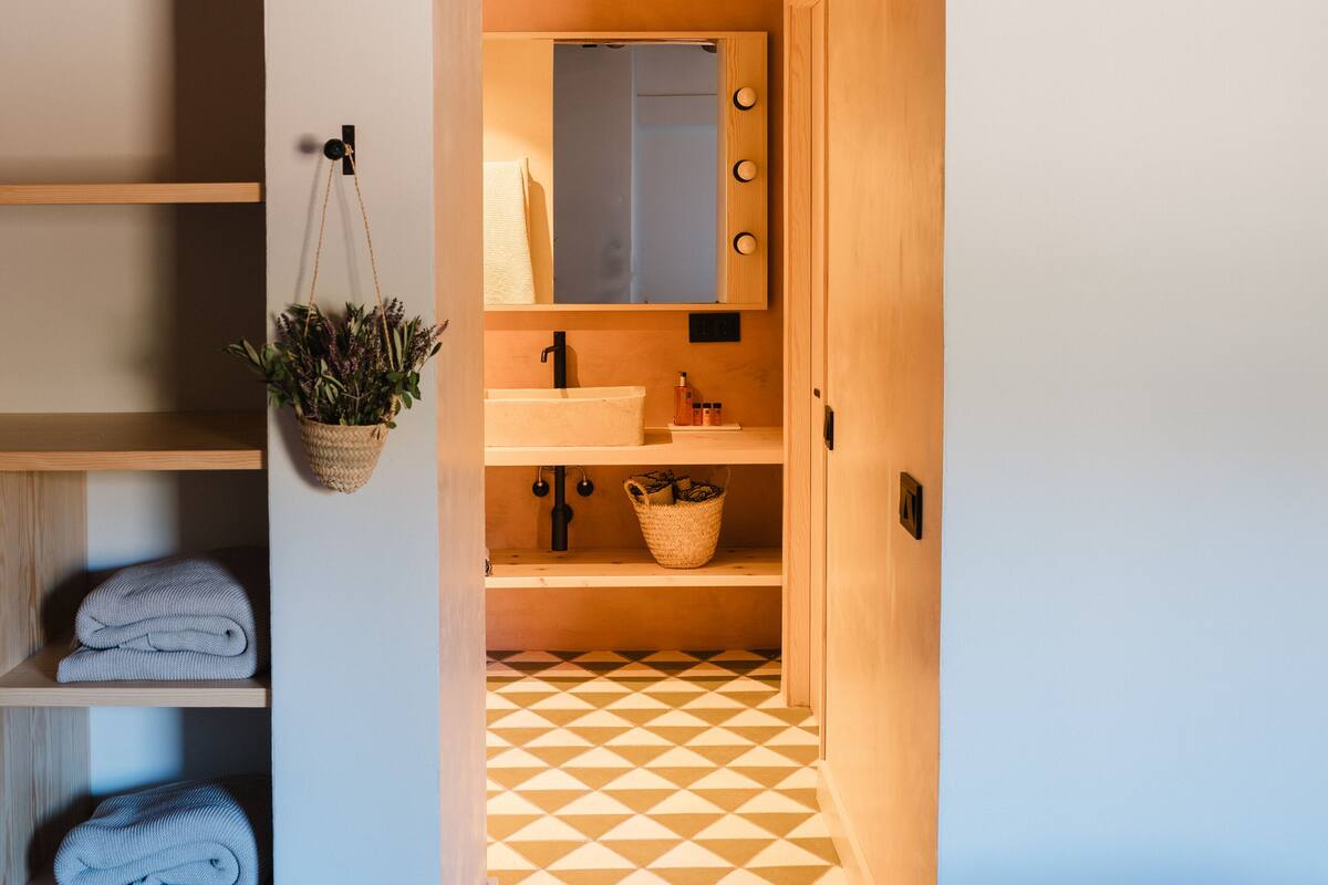 A narrow hallway leading to a bathroom with wooden beams, a hanging plant, and a patterned floor.