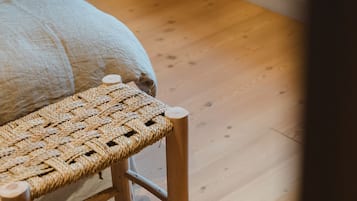 Un banco de madera con asiento y respaldo trenzado, colocado sobre un suelo de madera junto a una cama con cabecera blanca.