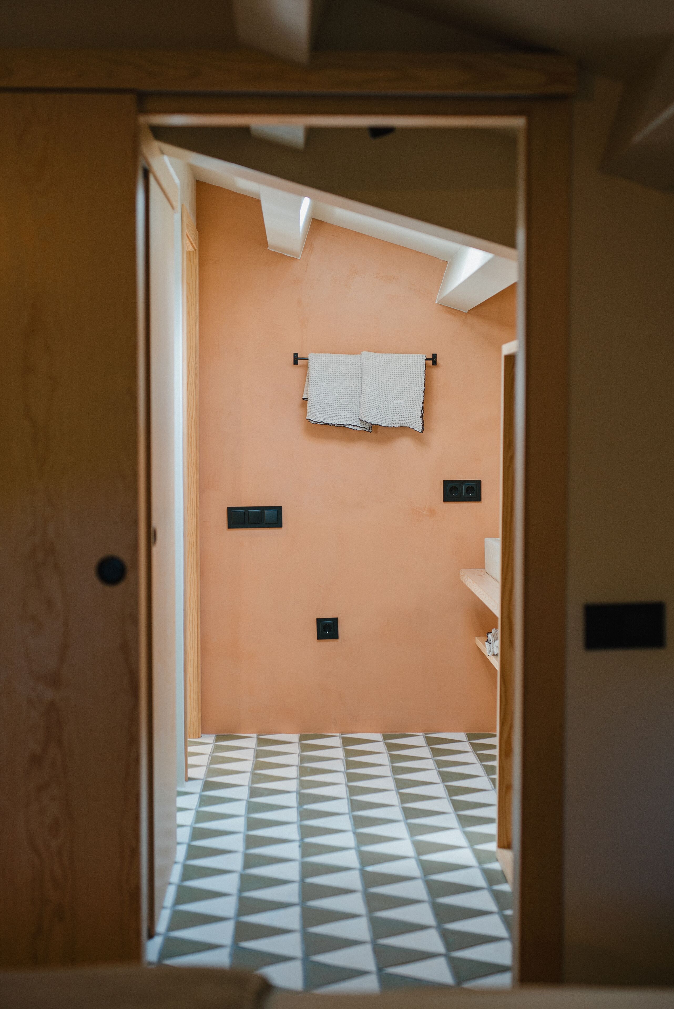 Un pasillo estrecho que conduce a un baño con piso estampado, una toalla colgada en la pared y un estante con objetos.