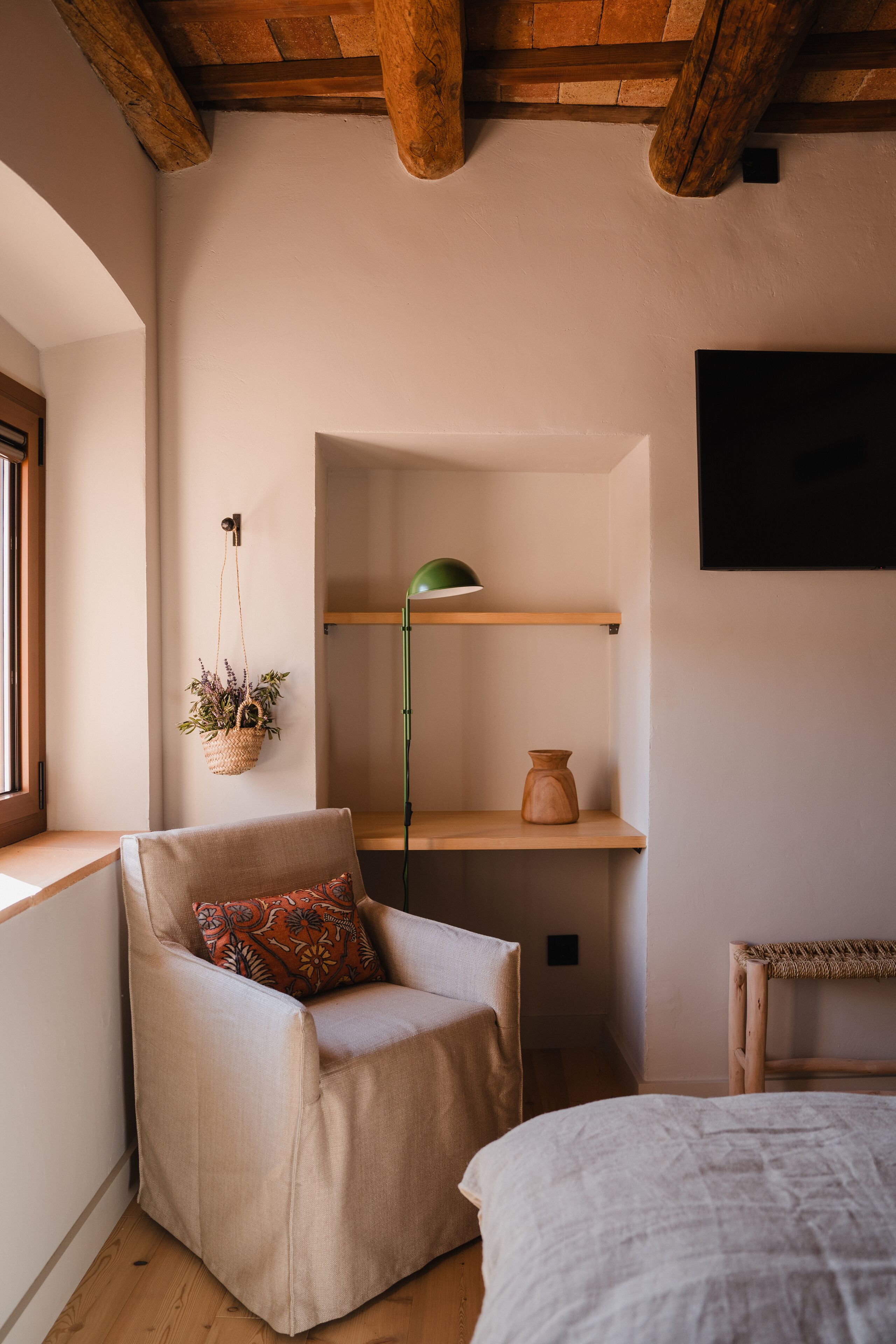 Una habitación acogedora con techo de madera, una silla con cojín, una mesita de noche, una planta colgante y una estantería con una lámpara y un jarrón.
