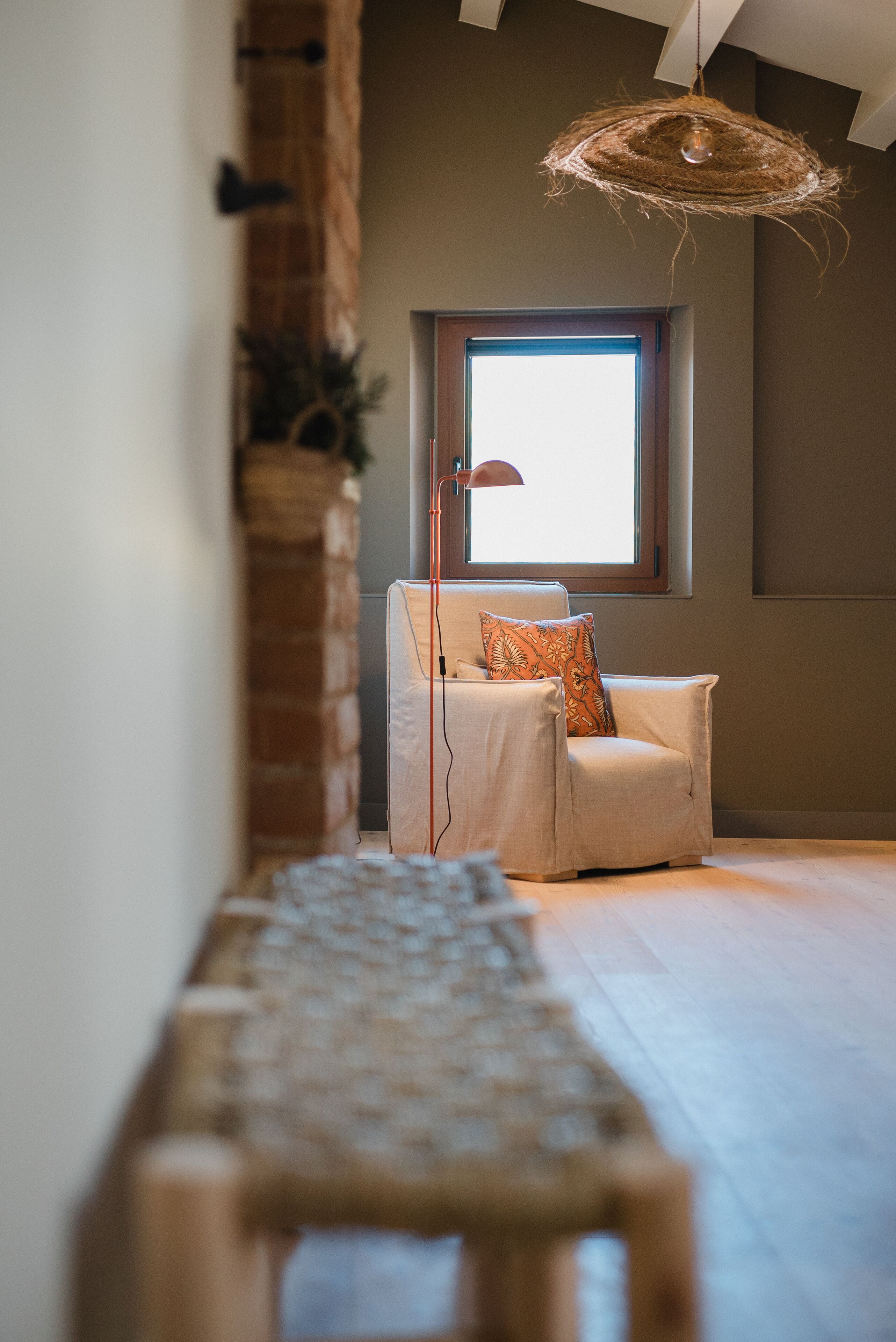 Un salon moderne avec un canapé blanc, un coussin décoratif à motifs, un luminaire suspendu en fibres tressées et une grande fenêtre laissant entrer la lumière naturelle.