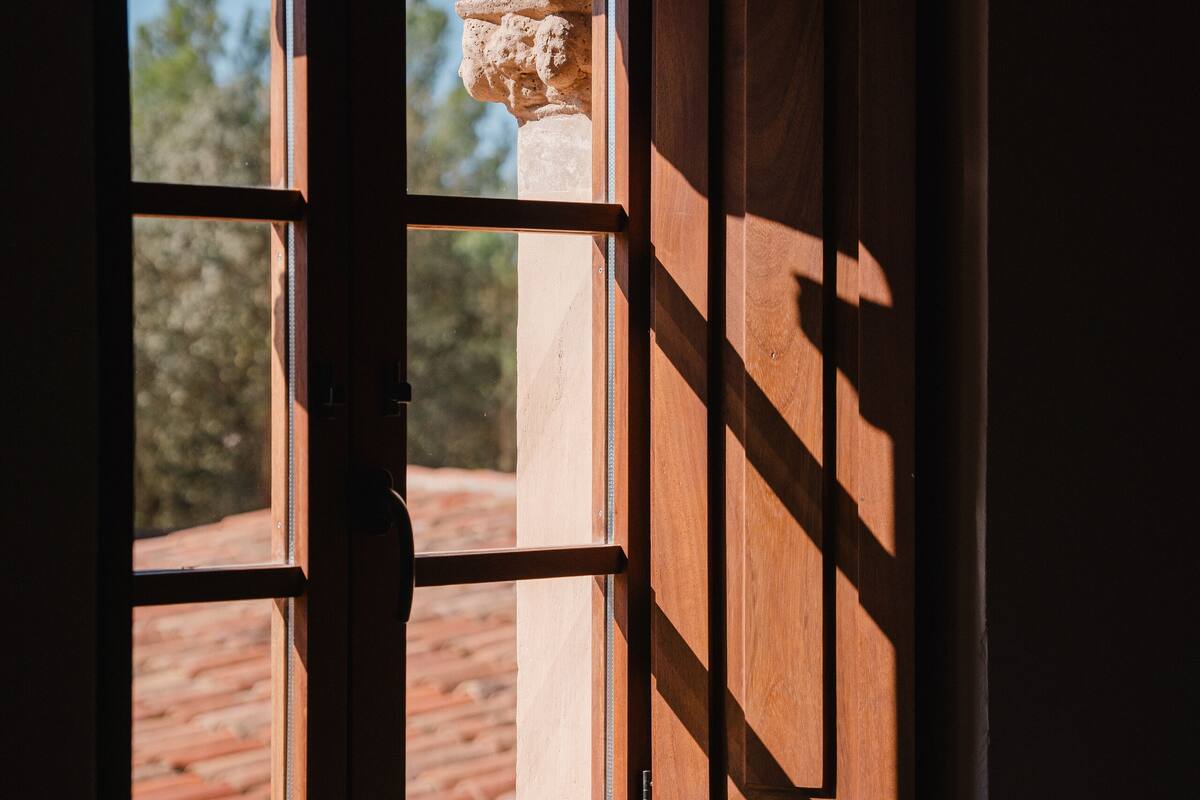 A window with wooden shutters, a view of a red-tiled roof, and a stone structure outside.