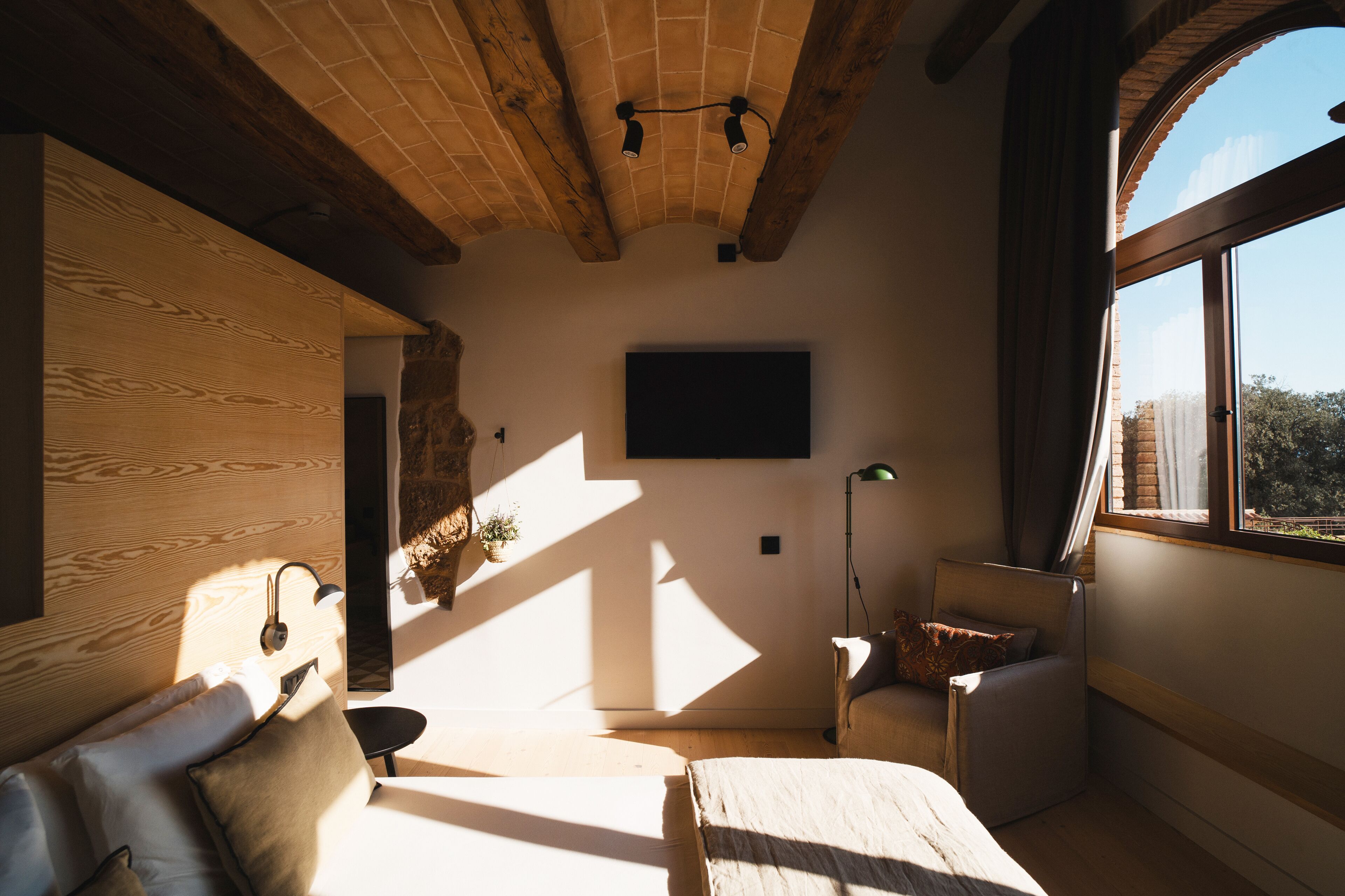 A room with a wooden ceiling, a bed, a chair, a desk, and a large window.