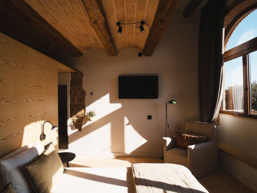 A room with a wooden ceiling, a bed, a chair, a desk, and a large window.
