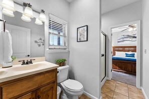 Ein Badezimmer mit Toilette, einem Waschbecken mit Holzschrank, einem Fenster mit Jalousien und einer Tür zu einem Schlafzimmer.