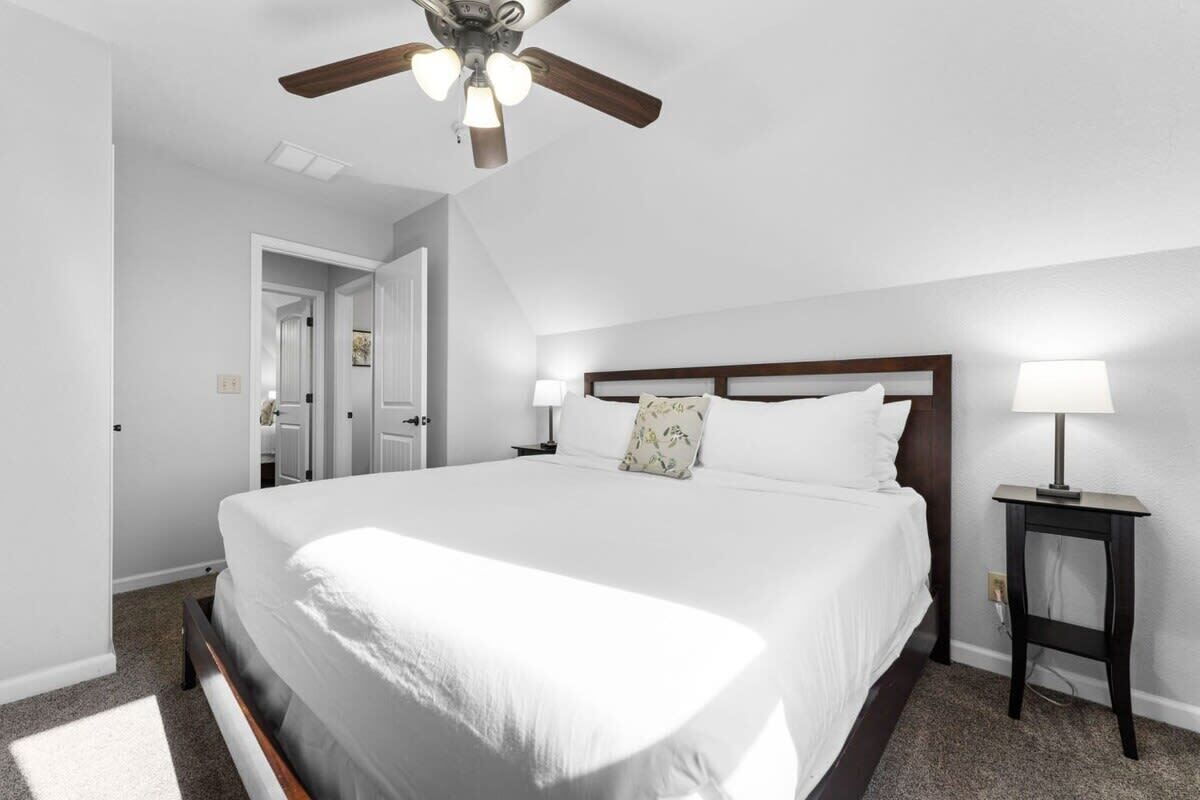 A bedroom with a large bed, a ceiling fan, a nightstand with a lamp, and a door leading to another room.