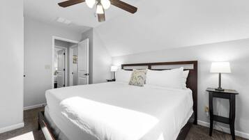 A bedroom with a large bed, a ceiling fan, a nightstand with a lamp, and a door leading to another room.
