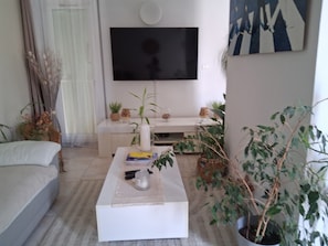 A living room with a flat-screen TV mounted on the wall, a white sofa, a coffee table with books and a vase, and several potted plants.