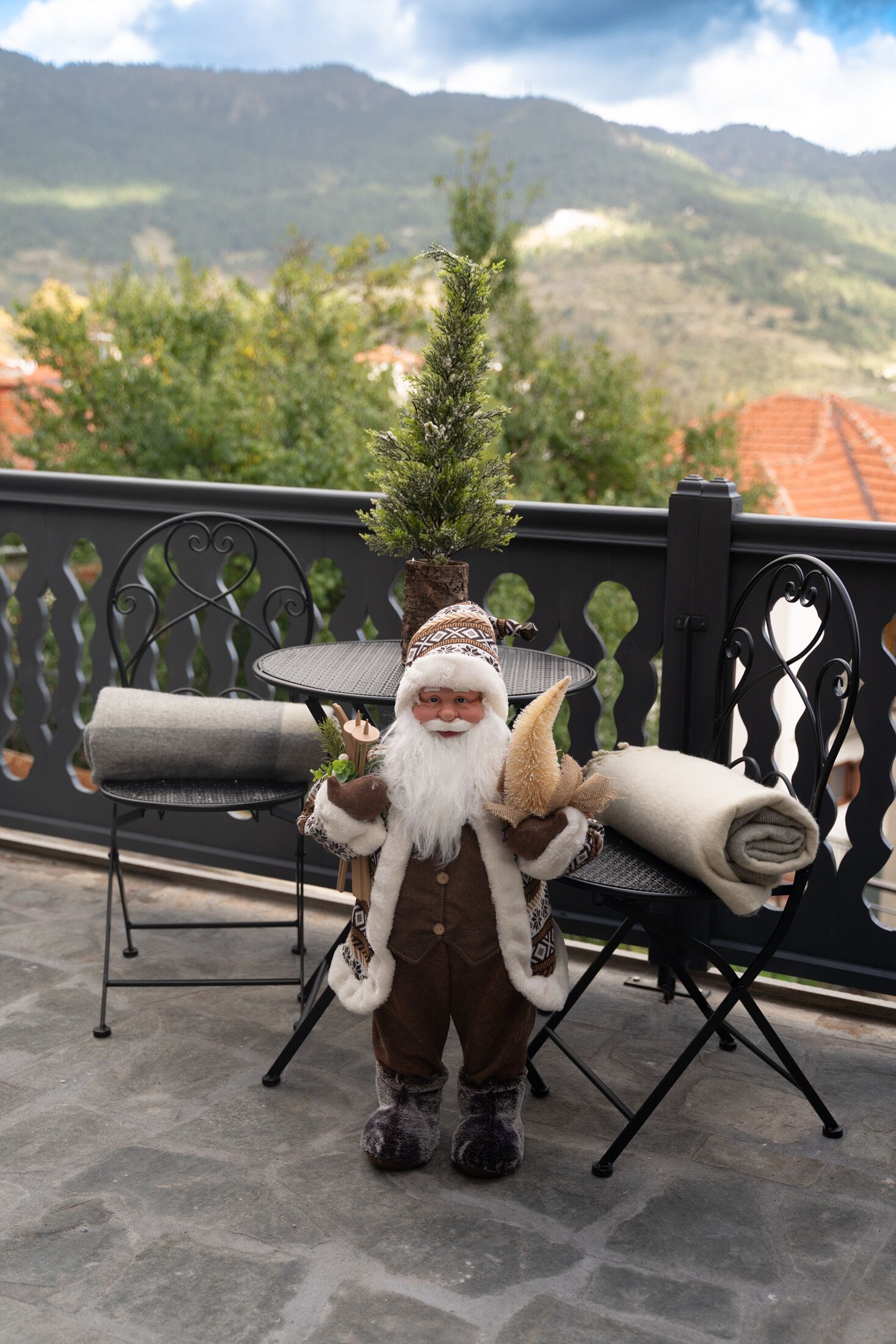 A Santa Claus figure on a balcony with a view of mountains and a tiled roof in the background.