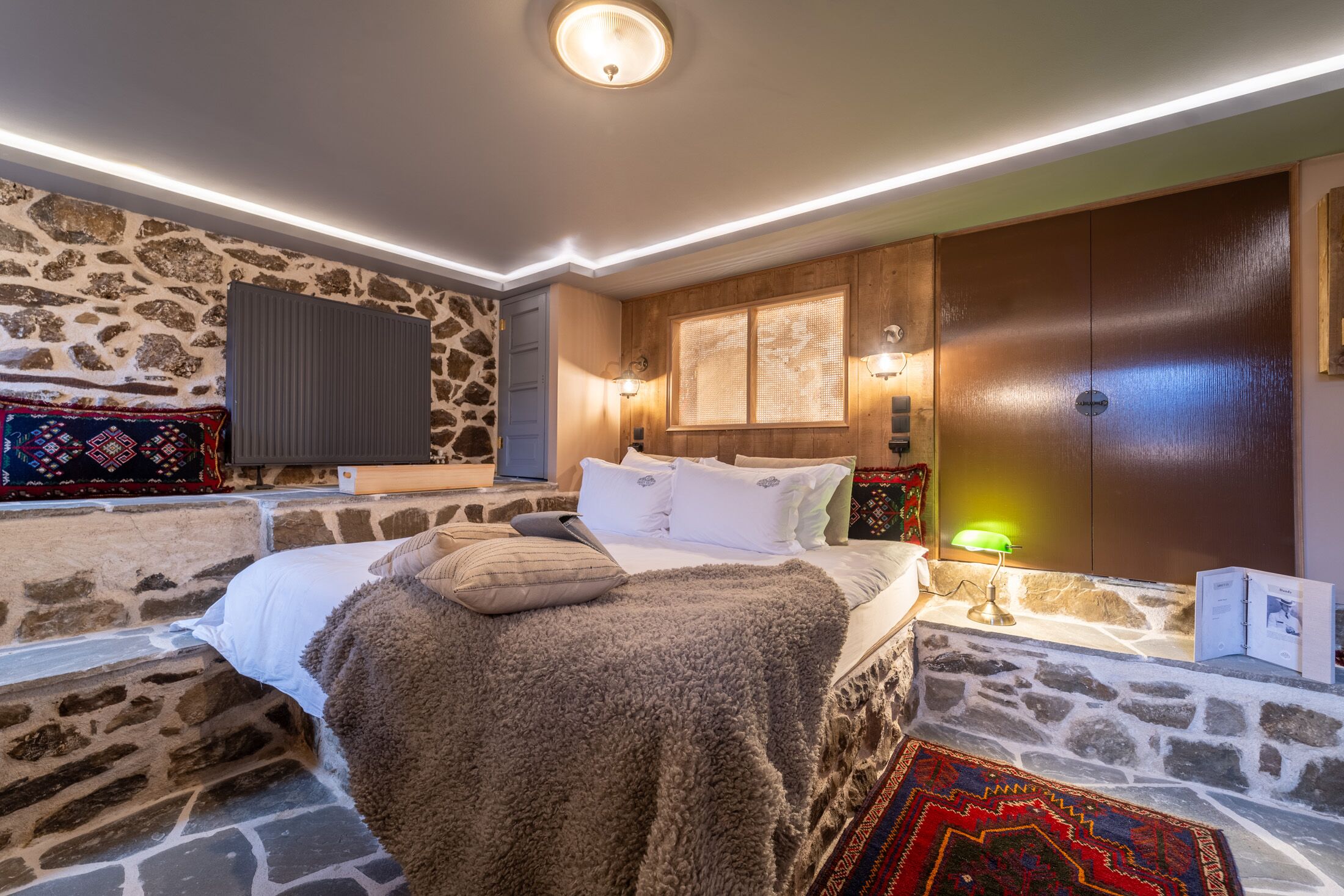 Un dormitorio con pared de piedra, una cama con manta mullida, un armario de madera y un televisor montado.