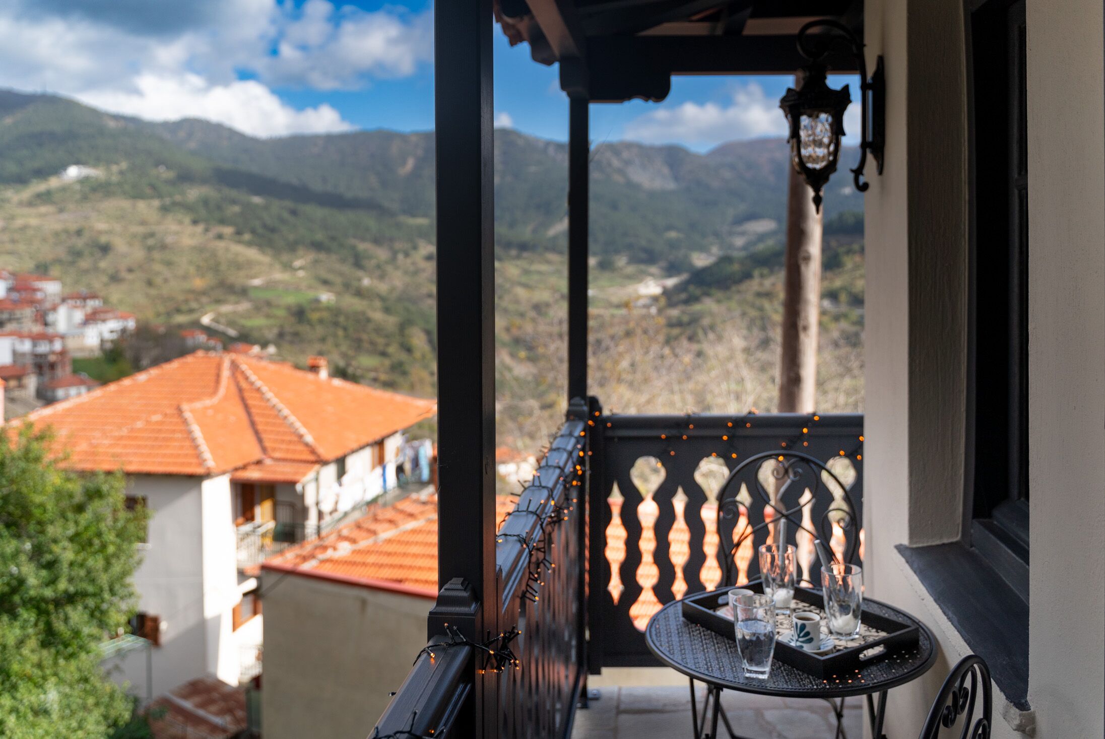 A balcony with a table and chairs, overlooking a mountainous landscape with buildings in the distance.