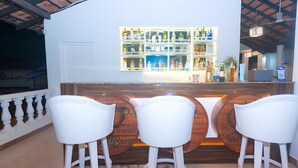 A bar area with white bar stools and wooden barrels, a white wall with a shelf of bottles, and a ceiling fan. - Lillywoods NV beach Resort (Calangute)