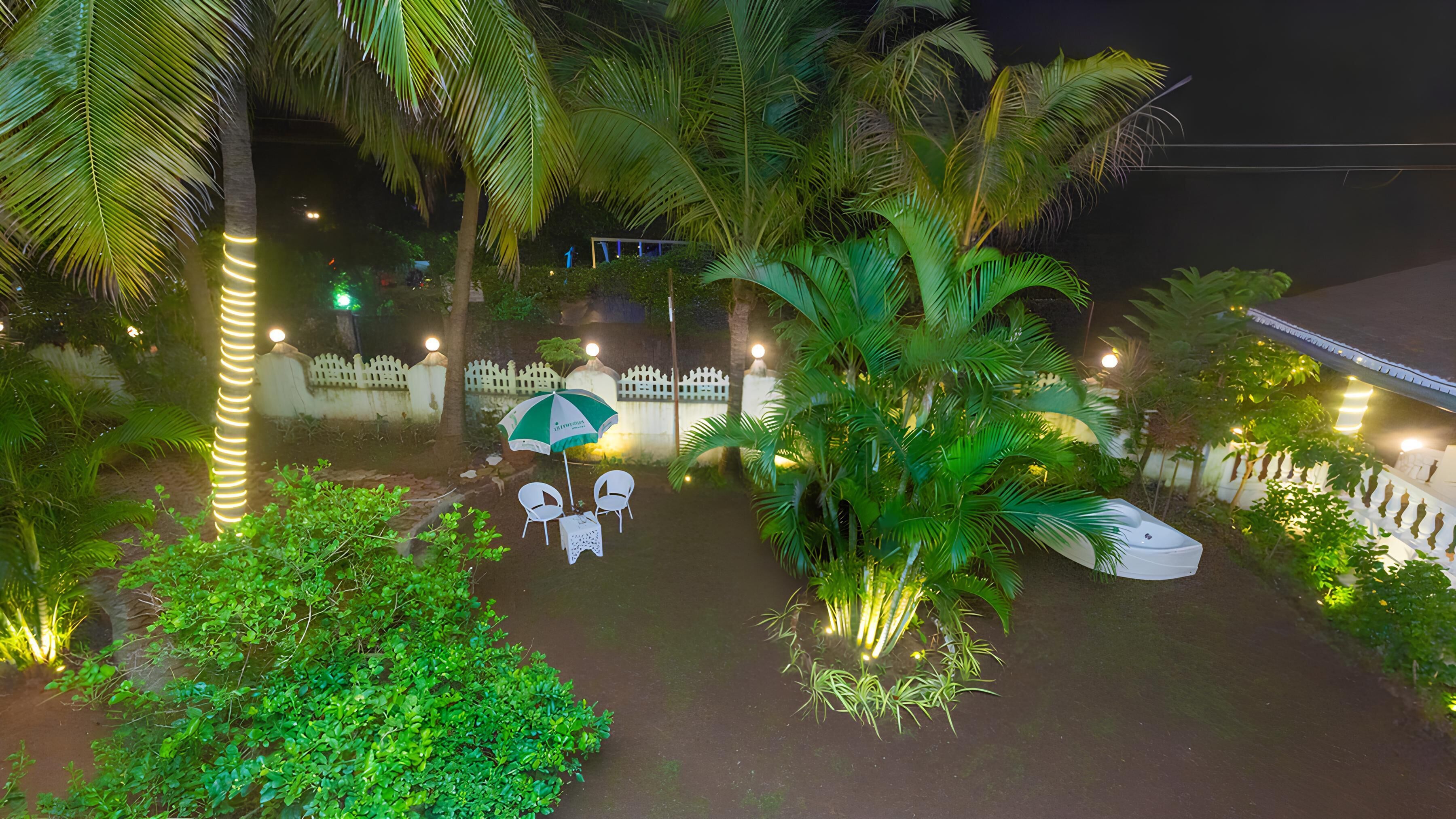 A nighttime outdoor seating area with tables and chairs, surrounded by lush greenery and palm trees.
