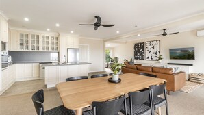 A modern kitchen with white cabinets and a central island, a dining area with a wooden table and black chairs, and a living area with a brown leather sofa and abstract wall art. - Como - Absolute Beachfront with Uninterrupted Convent Beach Views - Holiday Yamba (Yamba)