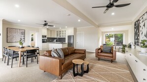 A spacious living area with a brown leather sofa, a dining table with chairs, a kitchen with white cabinets, and a ceiling fan. - Como - Absolute Beachfront with Uninterrupted Convent Beach Views - Holiday Yamba (Yamba)