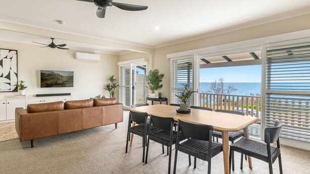 A spacious living room with a brown leather sofa, a wooden dining table with six chairs, and a large window with a view of the sea. - Como - Absolute Beachfront with Uninterrupted Convent Beach Views - Holiday Yamba (Yamba)