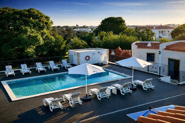 A rooftop pool with lounge chairs and umbrellas, surrounded by trees and buildings.
