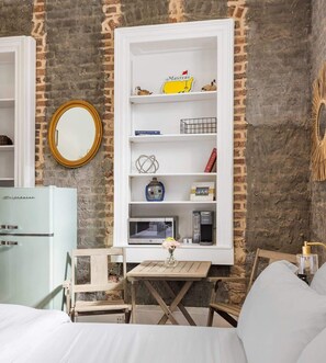 A compact hotel room with a brick wall, a white wardrobe with shelves, a small table with a vase, and a bed with white bedding. - Luxury French Quarter 1bd Suite w. Parking (Charleston)