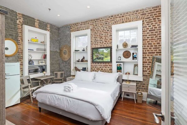 A bedroom with a brick wall, a bed, a desk, and shelves with various items.