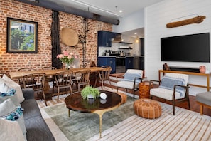 A modern living room with a brick wall, dining area, kitchen, and a large flat-screen TV.
