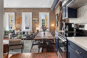 A spacious kitchen with a brick wall, a dining area with a wooden table and chairs, and a view through large windows.