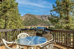 Eine Terrasse mit Glastisch und Stühlen, mit Blick auf eine gebirgige Landschaft und einem Grill.