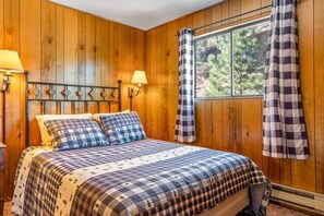 Ein Zimmer mit Holzwänden, einem Bett, zwei Wandlampen und einem Fenster mit karierten Vorhängen.